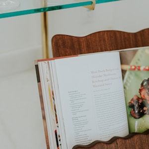 May include: A wooden cookbook stand holds an open cookbook with a recipe titled "Mini Duck Burgers with Shiitake Mushrooms, Ketchup and Chinese Mustard Sauce." The stand is made of dark wood and has a wavy top edge. A glass shelf and a bowl with fruit are in the background.