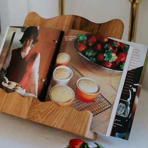 May include: A wooden cookbook stand holds an open cookbook with images of baked goods and strawberries. The stand is on a white surface, with a bowl of fresh strawberries in the foreground. Glassware is visible in the background.