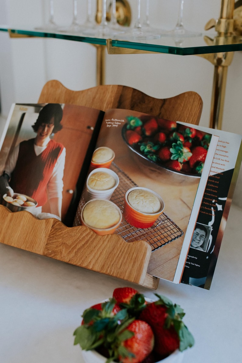 May include: A wooden cookbook stand holds an open cookbook with images of baked goods and strawberries. The stand is on a white surface, with a bowl of fresh strawberries in the foreground. Glassware is visible in the background.