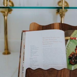 May include: A wooden cookbook stand holds an open cookbook with a recipe for Mini Duck Burgers. The stand has a wavy edge design. The background includes a glass shelf and gold accents.