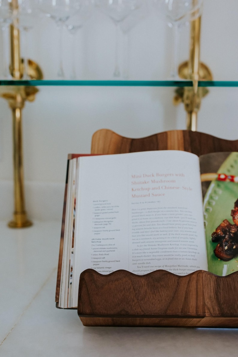 May include: A wooden cookbook stand holds an open cookbook with a recipe for Mini Duck Burgers. The stand has a wavy edge design. The background includes a glass shelf and gold accents.