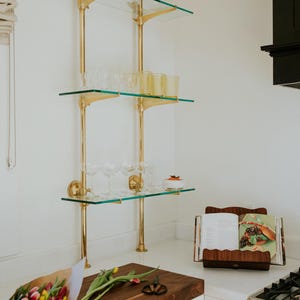 May include: A kitchen scene featuring a three-tiered glass shelf with brass supports, holding glassware. A wooden cutting board with tulips and a cookbook are on the countertop. The kitchen has white cabinets and a black stove.
