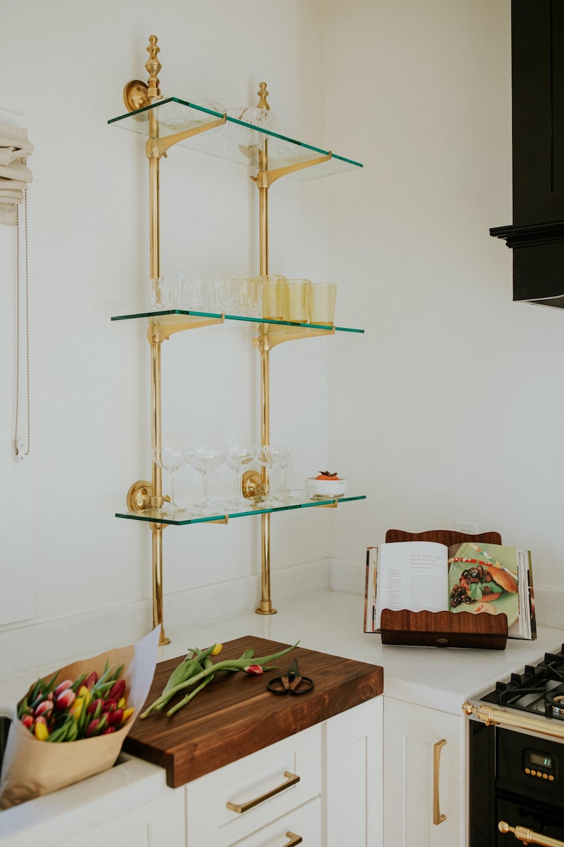May include: A kitchen scene featuring a three-tiered glass shelf with brass supports, holding glassware. A wooden cutting board with tulips and a cookbook are on the countertop. The kitchen has white cabinets and a black stove.