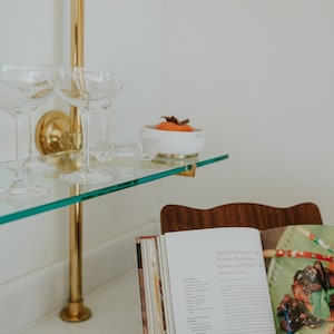 May include: A glass shelf with champagne glasses, a small white bowl with an orange fruit, and a wooden cookbook stand holding an open book. The shelf is supported by a gold-colored metal rod.