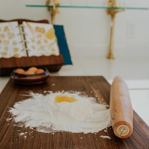May include: A wooden cutting board with flour and an egg yolk, ready for pasta making. A wooden rolling pin rests on the board. A cookbook and a bowl of eggs are in the background, suggesting a culinary theme.