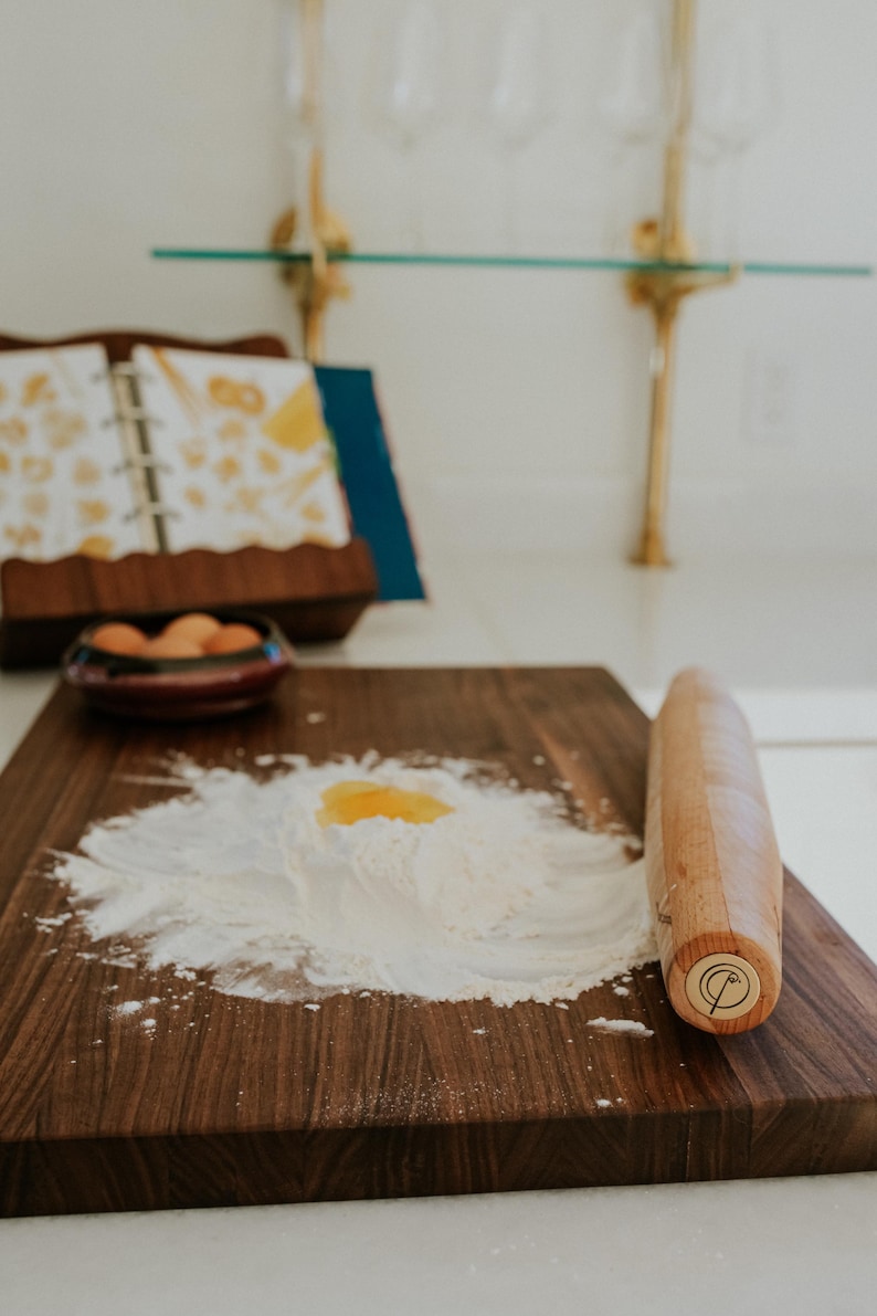 May include: A wooden cutting board with flour and an egg yolk, ready for pasta making. A wooden rolling pin rests on the board. A cookbook and a bowl of eggs are in the background, suggesting a culinary theme.