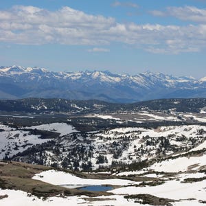 May include: A high-altitude landscape featuring a small, dark blue alpine lake surrounded by patches of white snow and dark green coniferous trees, with a vast range of snow-capped mountain peaks under a bright blue sky with scattered white clouds.