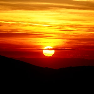 Puede incluir: Una vibrante puesta de sol naranja y amarilla con un gran sol redondo poniéndose detrás de una oscura cordillera montañosa.