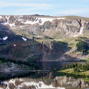 May include: A mountain lake with a reflection of the snow-capped mountains in the water. The lake is surrounded by green trees and hills.