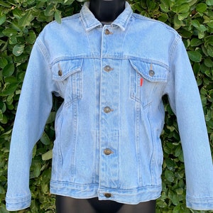 May include: Light blue denim jacket with button closures, two chest pockets, and two side pockets. The jacket has a classic design with a button-down front and long sleeves. A small red tag is visible on the front pocket. The jacket is displayed on a black mannequin.