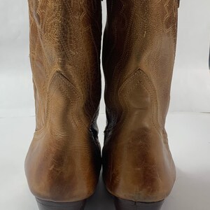 May include: A pair of brown leather cowboy boots. The boots feature a pointed toe, decorative stitching, and a stacked heel. The leather has a distressed look, and the boots are set against a white backdrop.