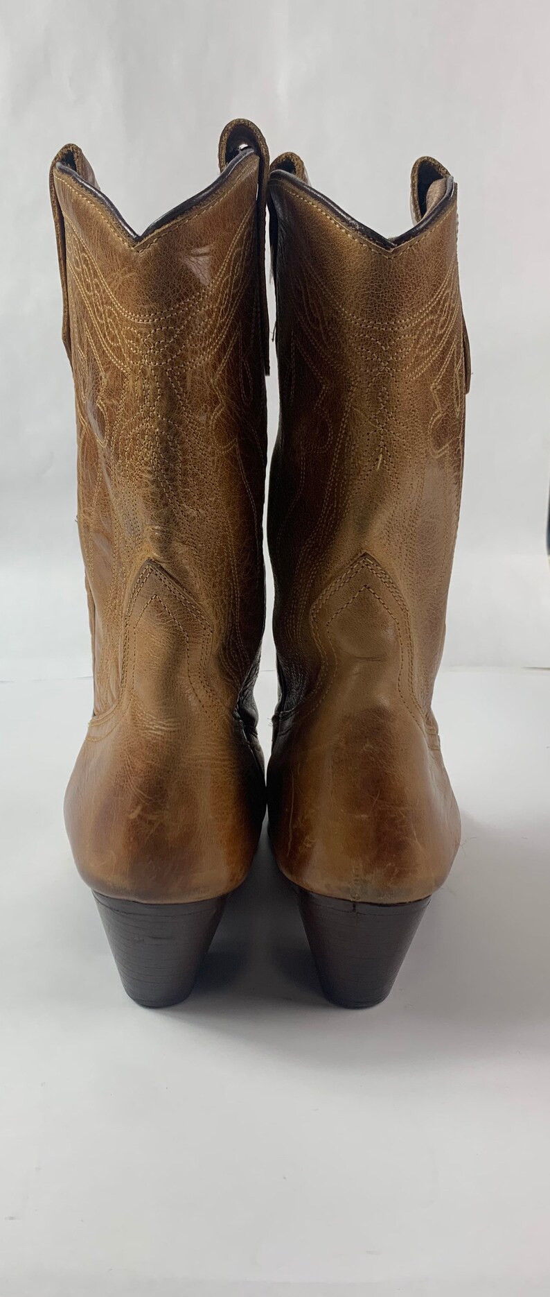 May include: A pair of brown leather cowboy boots. The boots feature a pointed toe, decorative stitching, and a stacked heel. The leather has a distressed look, and the boots are set against a white backdrop.