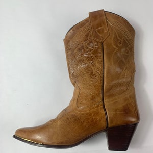 May include: A tan leather cowboy boot with decorative stitching and a pointed toe. The boot has a stacked heel and a gold-coloured metal accent on the toe. The boot is a medium height and is designed for wear.