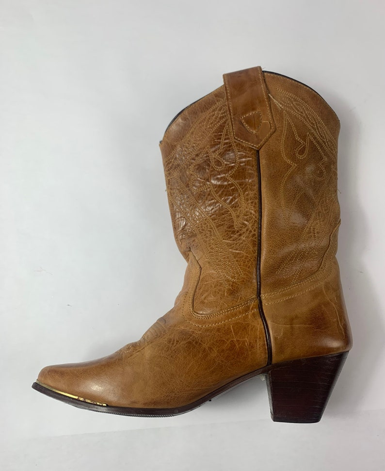 May include: A tan leather cowboy boot with decorative stitching and a pointed toe. The boot has a stacked heel and a gold-coloured metal accent on the toe. The boot is a medium height and is designed for wear.
