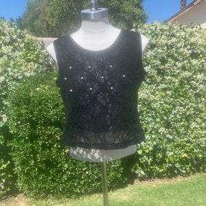 May include: A black sleeveless top made of lace fabric, adorned with small, round, white pearls. The top is displayed on a mannequin against a backdrop of green foliage and a sunny sky. The top has a scoop neckline.