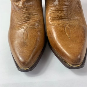 May include: Close-up of a pair of brown leather cowboy boots. The boots have pointed toes, decorative stitching, and a distressed finish. The soles have a dark brown colour with a gold trim.