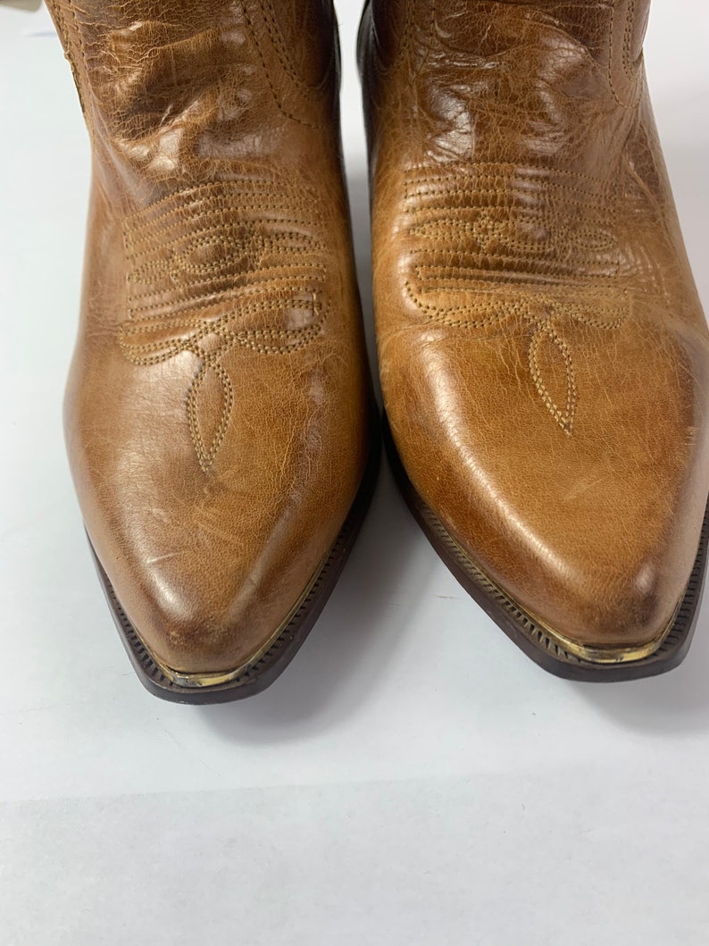 May include: Close-up of a pair of brown leather cowboy boots. The boots have pointed toes, decorative stitching, and a distressed finish. The soles have a dark brown colour with a gold trim.
