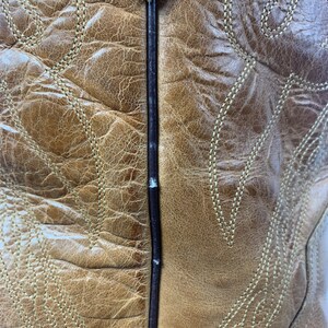 May include: Close-up of a tan leather cowboy boot. The boot features intricate stitching in a lighter tan colour, creating decorative patterns. The leather has a distressed appearance, with visible texture and colour variations.