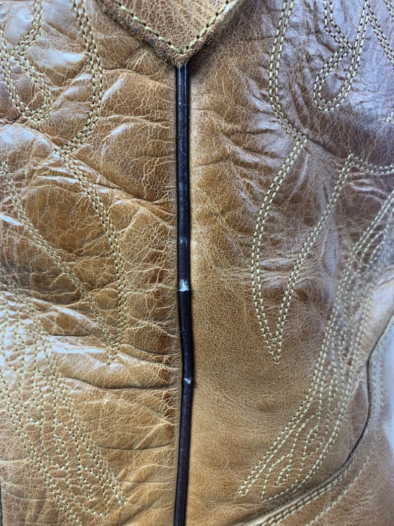 May include: Close-up of a tan leather cowboy boot. The boot features intricate stitching in a lighter tan colour, creating decorative patterns. The leather has a distressed appearance, with visible texture and colour variations.
