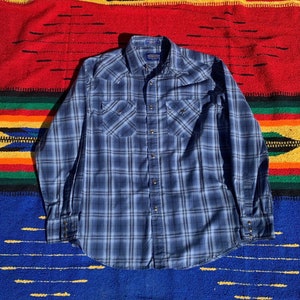 May include: Blue plaid button-down shirt with two chest pockets and snap closures. The shirt features a classic Western style with a pointed collar and long sleeves. The shirt is displayed on a colorful woven blanket.