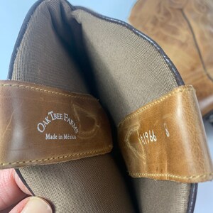 May include: Close-up of a pair of light brown leather cowboy boots. The interior of the boot is visible, showing a tan lining and a leather strap with the text "Oak Tree Farms Made in Mexico". The size is 8.