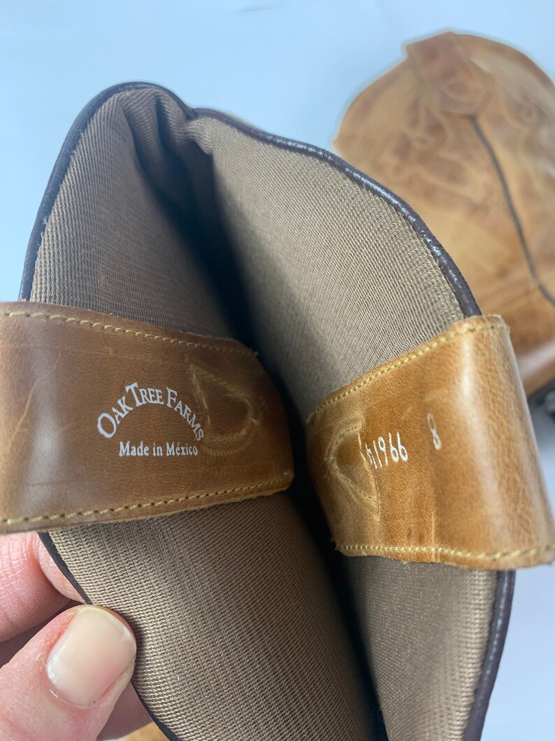 May include: Close-up of a pair of light brown leather cowboy boots. The interior of the boot is visible, showing a tan lining and a leather strap with the text "Oak Tree Farms Made in Mexico". The size is 8.
