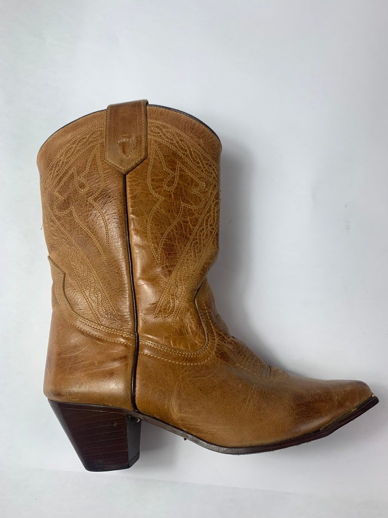 May include: A single tan leather cowboy boot with decorative stitching. The boot has a pointed toe and a dark brown heel. The leather appears worn, giving it a vintage look. The boot is set against a white background.