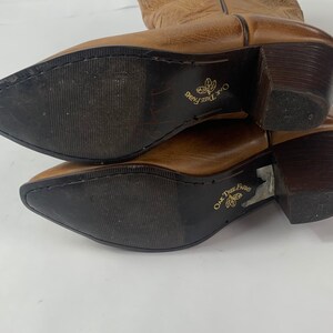 May include: A pair of brown leather cowboy boots with pointed toes and dark brown heels. The soles are dark brown with the text "Oak Tree Farms" printed in gold. The boots are angled towards the viewer.
