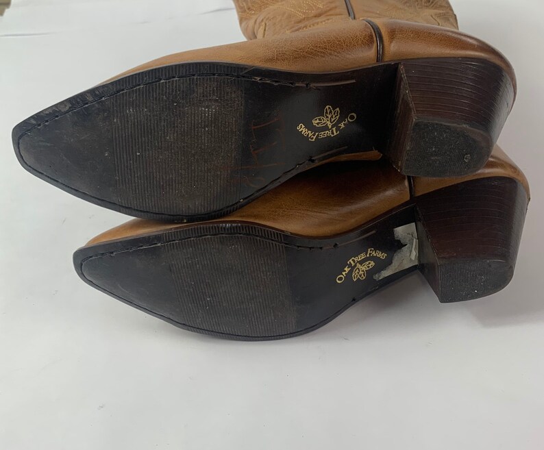 May include: A pair of brown leather cowboy boots with pointed toes and dark brown heels. The soles are dark brown with the text "Oak Tree Farms" printed in gold. The boots are angled towards the viewer.