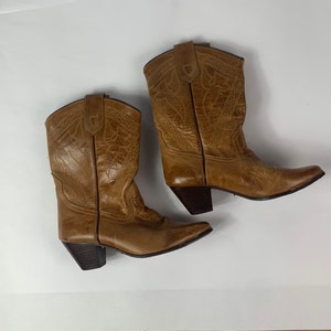 May include: A pair of brown leather cowboy boots. The boots have a pointed toe, a mid-height heel, and decorative stitching on the shaft. The boots are set against a white background.