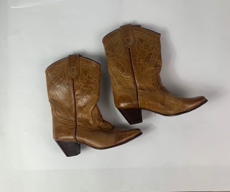 May include: A pair of brown leather cowboy boots. The boots have a pointed toe, a mid-height heel, and decorative stitching on the shaft. The boots are set against a white background.