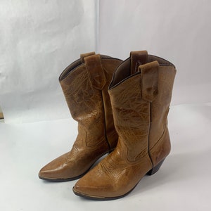 May include: A pair of brown cowboy boots with pointed toes and decorative stitching. The boots have a worn, textured appearance and a small heel. The boots are made of leather and have a classic Western style.
