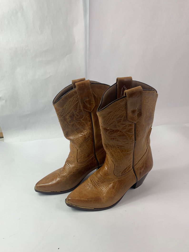 May include: A pair of brown cowboy boots with pointed toes and decorative stitching. The boots have a worn, textured appearance and a small heel. The boots are made of leather and have a classic Western style.