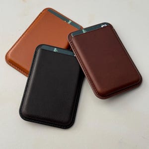 May include: Three rectangular card holders in tan, dark brown, and black leather, each featuring rounded corners and visible stitching along the edges. Each holder contains a single card partially inserted into the top slot, displaying a dark green edge.