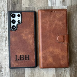 Puede incluir: Una funda de teléfono de cuero marrón con un borde negro y una solapa que se cierra con un botón. La funda tiene un monograma con las iniciales "LBH" en la parte delantera.