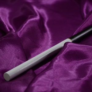 Può includere: Un oggetto a forma di bacchetta bianco e nero poggia su un tessuto di raso viola scuro. La bacchetta ha un'impugnatura bianca e una punta nera. Lo sfondo è di un viola ricco e strutturato, creando un contrasto sorprendente.