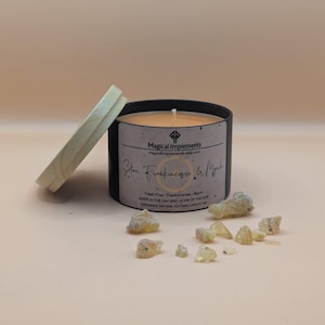 May include: A black candle jar with a wooden lid. The candle is labeled "Solar Frankincense & Myrrh" and "Magical Implements". The jar is surrounded by small pieces of amber-colored resin.