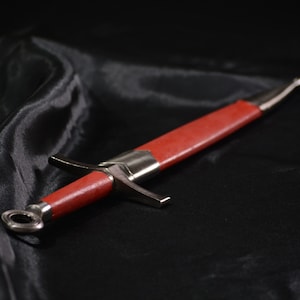 Puede incluir: Una daga roja y plateada con mango de cuero sobre un fondo de tela negra.