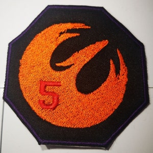 Könnte beinhalten: Ein orangefarbenes gesticktes Symbol der Rebellenallianz mit einer roten Nummer 5 in der Mitte, auf einem schwarzen achteckigen Patch mit einem violetten Rand.