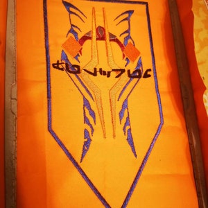 Könnte beinhalten: Ein orangefarbener Stoff-Patch mit einer blauen, gestickten Umrandung in Form einer Raute. Innerhalb der Raute befindet sich ein stilisiertes Design mit drei gelben Spitzen und einem blauen und orangefarbenen Muster. Der Text "CLO HUR7G6" ist in Gelb unterhalb des Designs gestickt.