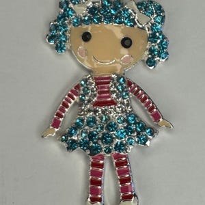 Puede incluir: Un colgante con forma de muñeca con acabado metálico plateado. La muñeca presenta un tono de piel claro, ojos negros y mejillas rosadas. El vestido y el pelo están adornados con pedrería de color turquesa. Los brazos y las piernas de la muñeca son rosas y blancos.