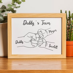 May include: A framed print with the text "Daddy's Team" and line art of three fists bumping. The names "Daddy", "Megan", and "Scarlett" are written below the fists. The frame is light brown.