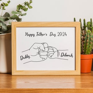 May include: A framed print with a light wood frame. The print features a black and white line drawing of two fists bumping, with the words "Happy Father's Day 2024" above and the names "Daddy" and "Deborah" below.