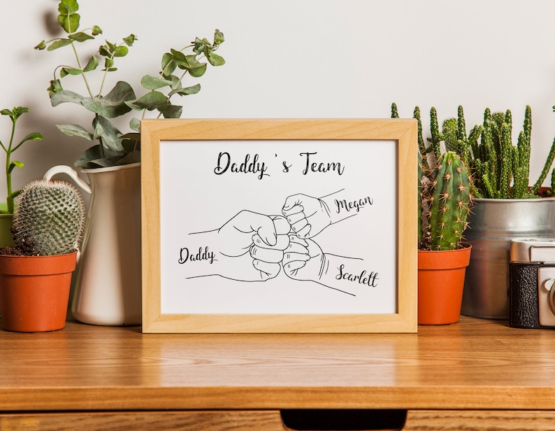 May include: A framed print with the text "Daddy's Team" and line art of three fists bumping. The names "Daddy", "Megan", and "Scarlett" are written below the fists. The frame is light brown.