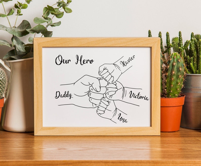 May include: A framed print with a light wood frame. The print features a black and white line drawing of hands in a fist bump, with the words "Our Hero" at the top and names "Daddy", "Xavier", "Victoria", and "Una" on the print.