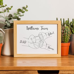 May include: A framed print with a light wood frame. The print features a black and white line drawing of four fists bumping, with the words "Williams Team" above and the names "Dad", "Gilbert", and "Norma" below.