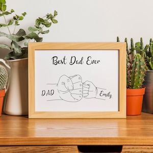 May include: A framed print with the text "Best Dad Ever" and the names "Dad" and "Emily" in black script. The artwork depicts two fists bumping. The frame is light brown, and the print is displayed on a wooden surface.