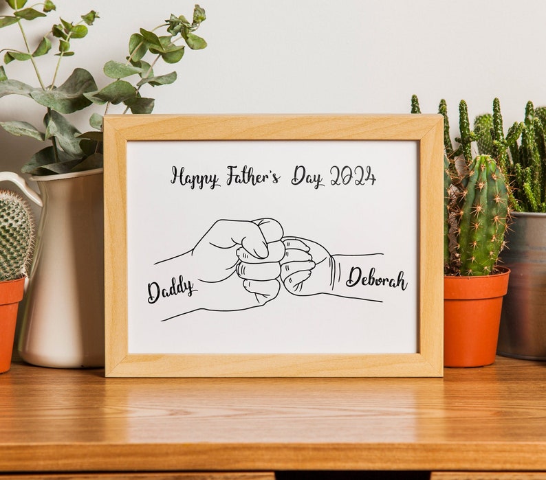 May include: A framed print with a light wood frame. The print features a black and white line drawing of two fists bumping, with the words "Happy Father's Day 2024" above and the names "Daddy" and "Deborah" below.