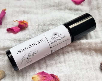 May include: A black roller bottle with a white label that reads "sandman. for Restful Sleep" and "INHALE Botanicals". The bottle is on a white fabric background with pink rose petals.