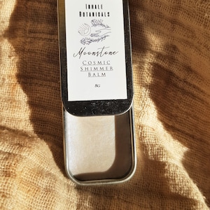 May include: A silver tin with a white label that reads "Inhale Botanicals Moonstone Cosmic Shimmer Balm 8g". The label has a graphic of a sun, a hand, and a flower.
